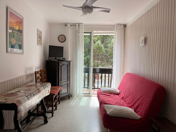 Una sala de estar con un sofá rojo, una mesa de madera con mantel estampado, un televisor en un mueble bajo y un balcón con vistas a árboles y un edificio.