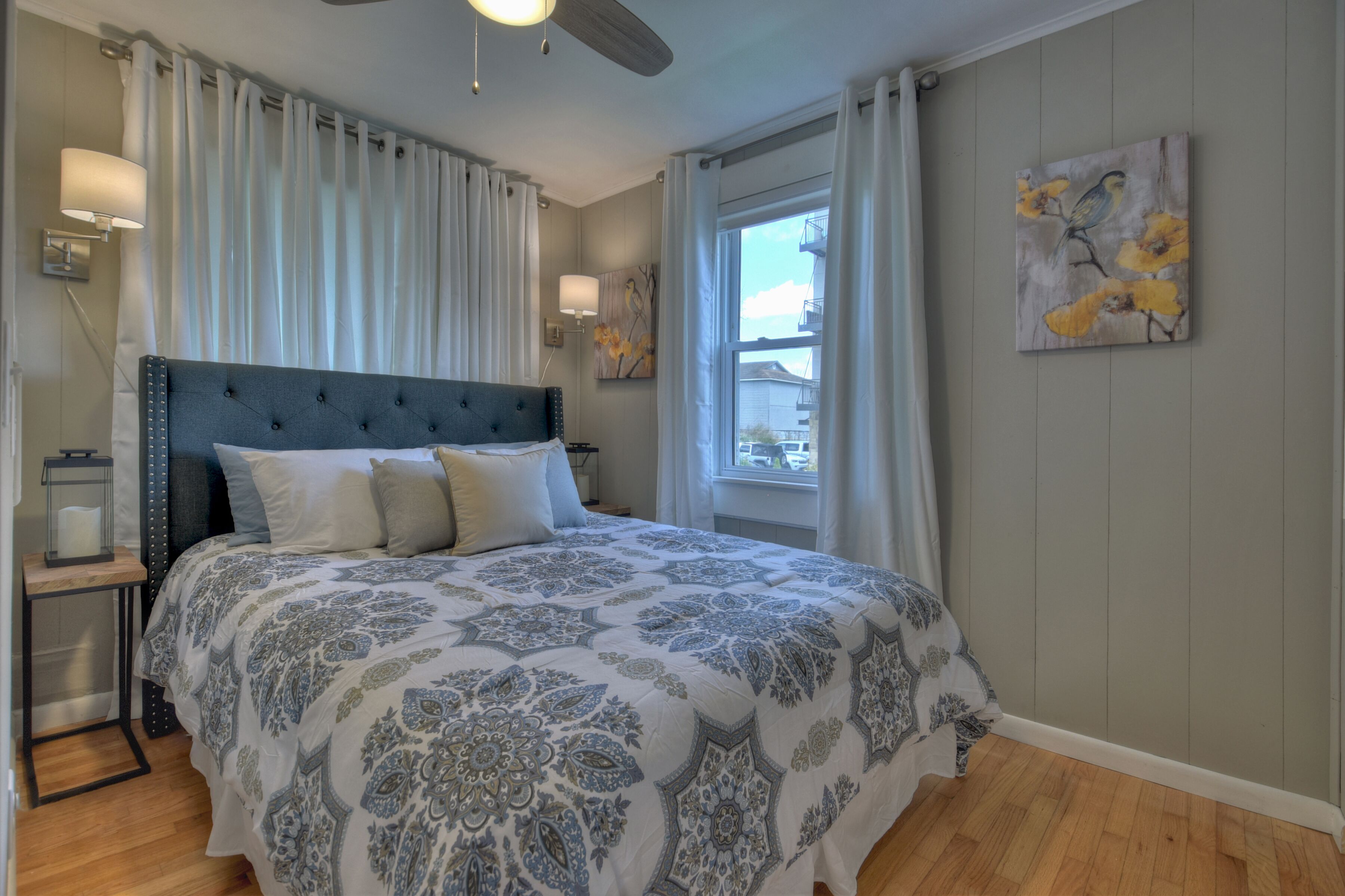 A bedroom with a large bed, a ceiling fan, a nightstand with a lamp, and a window with curtains.