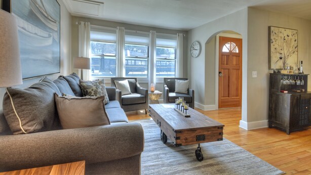 A living room with a gray sofa, a wooden coffee table, and a view of a train station through large windows. - Lovely Brick Cottage in Downtown Blue Ridge (Blue Ridge)