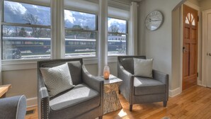 A room with two grey upholstered chairs with patterned cushions, a small wooden side table, and a large window with a view of a train passing by. - Lovely Brick Cottage in Downtown Blue Ridge (Blue Ridge)