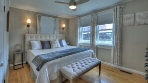 A bedroom with a large bed, a bench, two wall-mounted lamps, a ceiling fan, and a window with curtains. - Lovely Brick Cottage in Downtown Blue Ridge (Blue Ridge)