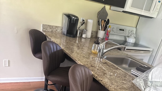 Una cocina con encimera de granito, pileta de acero inoxidable y microondas. Hay tres taburetes de bar con patas negras y asientos marrones.