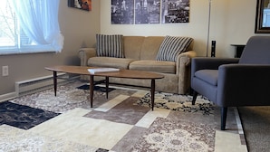Una sala de estar con sofá, sillón, una mesa de centro y alfombras estampadas. Se observan dos cuadros colgados en la pared y una lámpara de pie.