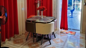 A dining area with a round table, chairs, and a fan. Red curtains and a white coat stand are visible.
