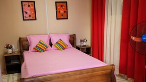 A bedroom with a wooden bed, pink bedding, red curtains, two wall art pieces, a nightstand with a lamp, and a patterned rug.