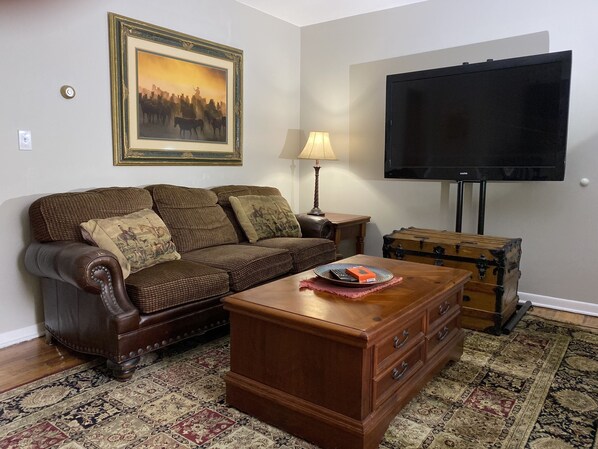 A living room with a brown leather sofa, a wooden coffee table, a patterned rug, a television, and a vintage suitcase.