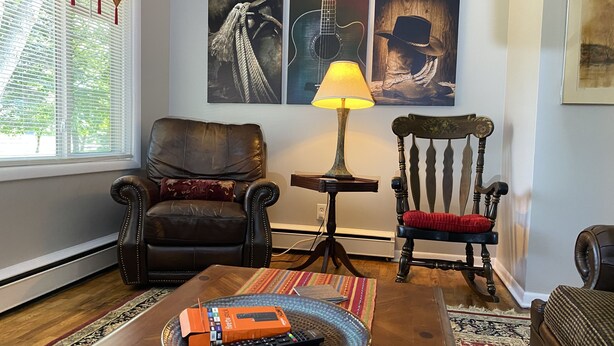 A living room with a leather armchair, a wooden chair, a coffee table with a remote control and a box, and artwork on the wall. - Fireside Ranch of Romeo (bruce twp)