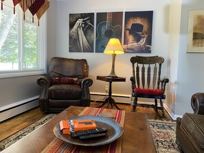 A living room with a leather armchair, a wooden chair, a coffee table with a remote control and a box, and artwork on the wall.