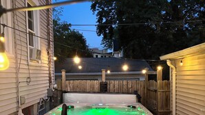 Jacuzzi en un patio trasero con valla de madera, luces colgantes y terraza de madera.