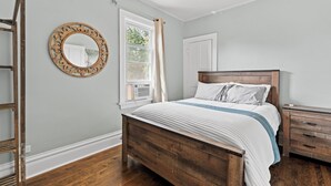 Un dormitorio con una cama de madera, un espejo redondo, un ventilador de techo, una ventana con cortinas y una mesita de noche de madera.