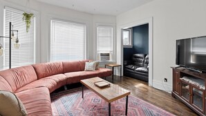 Una sala de estar moderna con un sofá seccional rosa, una mesa de centro de madera, una alfombra estampada, un sillón de cuero negro, un televisor sobre un mueble de madera y una planta colgante.