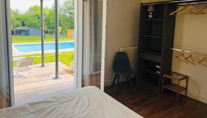 A bedroom with a large bed, a sliding glass door leading to a pool, a small table, and a dark-colored wardrobe.
