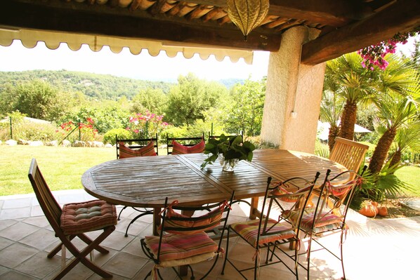 Une terrasse couverte avec une table ronde en bois et des chaises, entourée d’une végétation luxuriante et offrant une vue sur des collines lointaines.