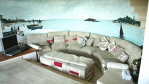 Ein Wohnzimmer mit einem großen Gemälde einer Küstenlandschaft, einem beigefarbenen Ecksofa, einem kleinen Tisch mit einem Laptop und einem Stauraum-Pouff.