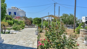 Un chemin de pierres mène à un bâtiment blanc aux volets bleus, entouré d’une végétation luxuriante et de plantes en fleurs.