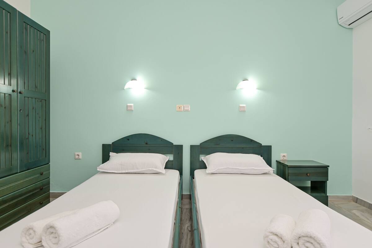 Habitación doble con dos camas individuales, un armario de madera, una mesita de noche e iluminación en la pared.