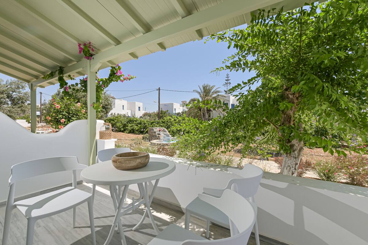 Un área de estar al aire libre con techo, con sillas y mesa blancas, con vista a un jardín con árboles y edificios al fondo.