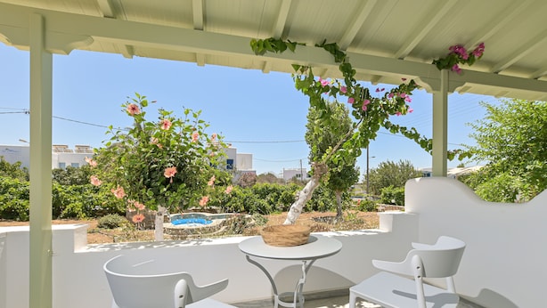 Un espace extérieur aménagé, abrité et doté de chaises blanches et d’une table, entouré de verdure et offrant une vue sur une piscine.