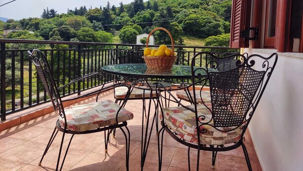 En balkon med et bord og stole, en kurv med citroner, og udsigt over et grønt landskab.