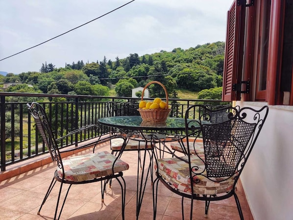 A balcony with a table and chairs, a basket of lemons, and a view of a green landscape.