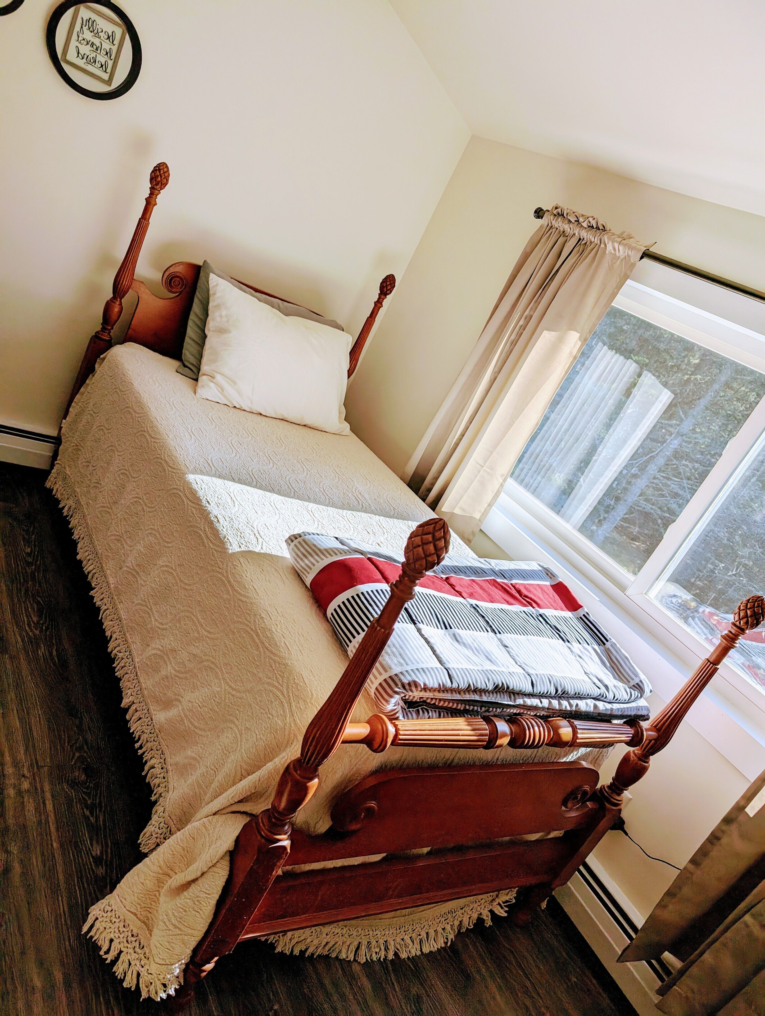 Una cama bien hecha con cabecera de madera, una ventana con cortinas y un cuadro enmarcado en la pared.