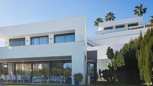 A modern two-story house with a flat roof, large windows, and a covered outdoor seating area.