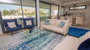 A modern living room with a glass coffee table, two armchairs, a sofa, and a kitchen area visible through glass doors.