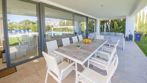 Een moderne buiten eethoek met een lange houten tafel en witte stoelen, met uitzicht op een zwembad en tuin.