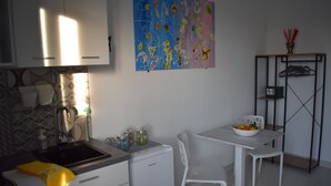 A modern kitchen with white cabinets, a sink, and a compact refrigerator. There's a dining area with a white table and chairs, and a colorful abstract painting on the wall.
