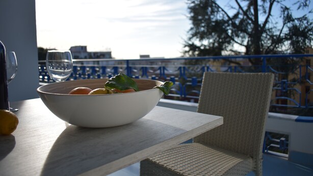 A table with a bowl of fruit and a glass of water on a balcony with a view of the sky and trees.