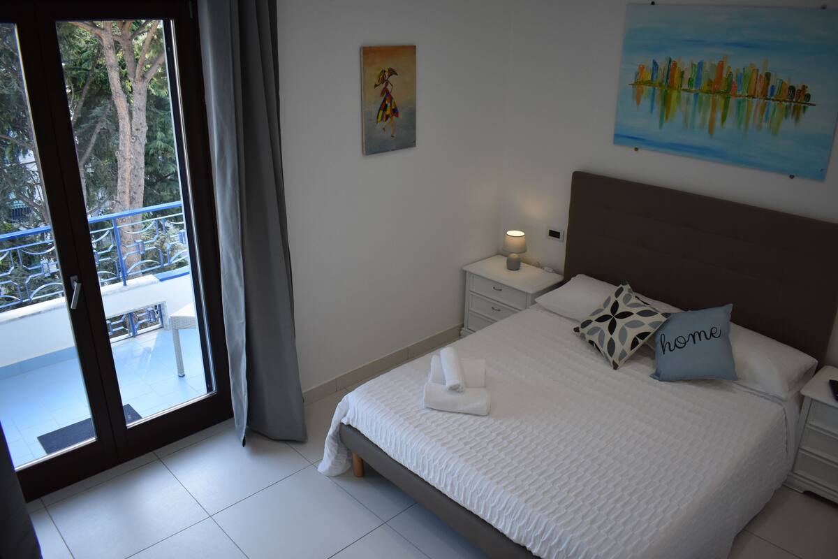 A bedroom with a bed, a nightstand, a lamp, a painting, and a view of a balcony.