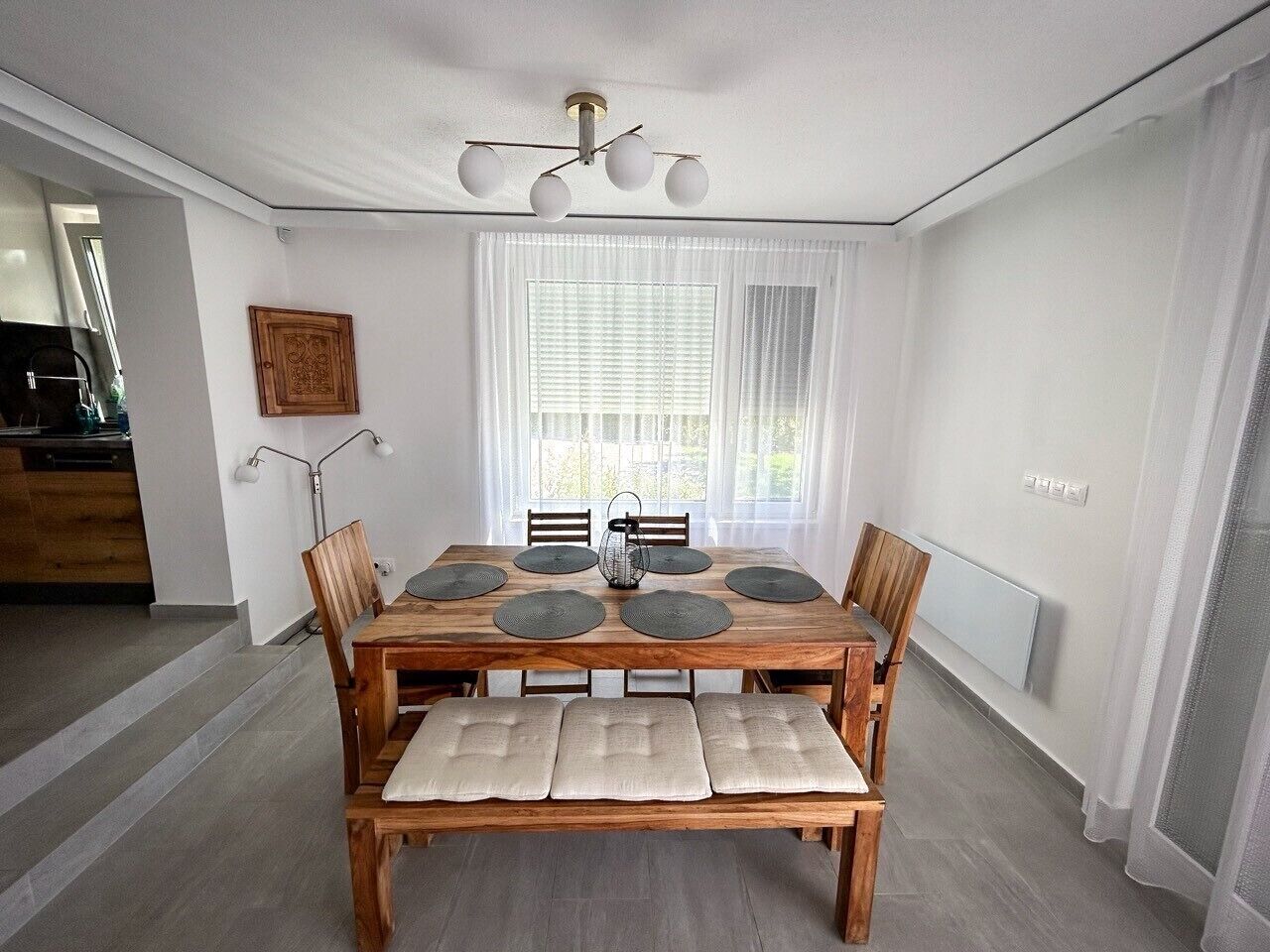 A dining area with a wooden table, six chairs, and a bench. There is a chandelier above the table, and a window with curtains behind it.