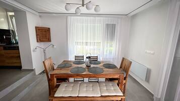 A dining area with a wooden table, six chairs, and a bench. There is a chandelier above the table, and a window with curtains behind it.
