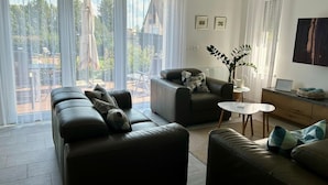 Un salon moderne avec un canapé, un fauteuil et une petite table d’appoint. De grandes fenêtres sont ornées de rideaux légers, et on y trouve une plante en pot ainsi qu’un tableau encadré accroché au mur.