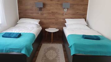 A room with two single beds, each with a teal blanket and pillow, a small round table, and two wall-mounted speakers.