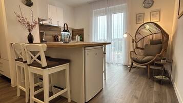 A compact kitchen with a dining area, a small refrigerator, and a wicker chair.