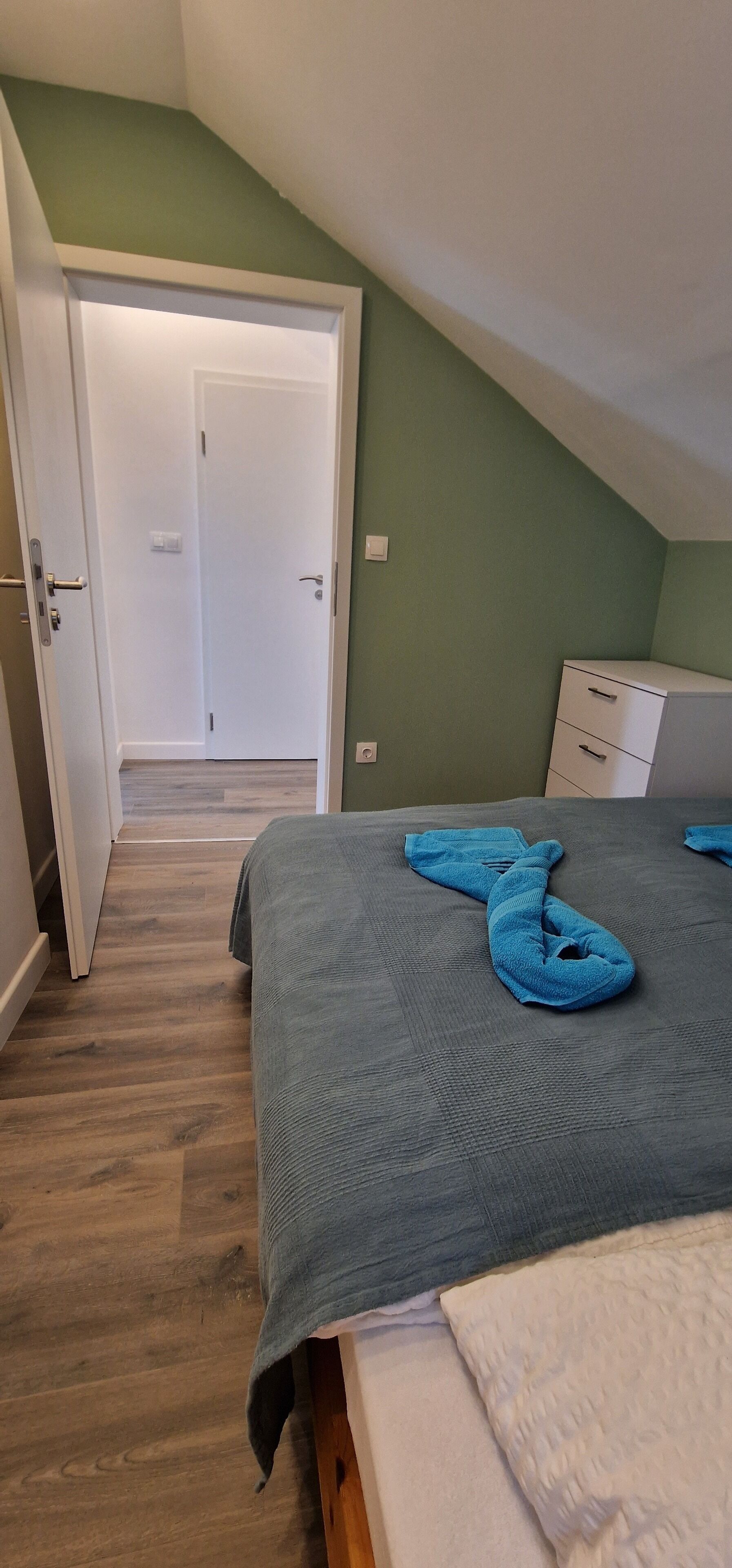 Une chambre avec un plafond en pente, un lit recouvert d’une couverture bleue, une commode blanche et une porte donnant sur une autre pièce.