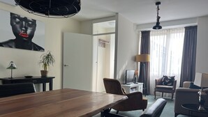 A modern hotel room with a wooden dining table, comfortable seating, a large window with curtains, and a flat-screen TV.