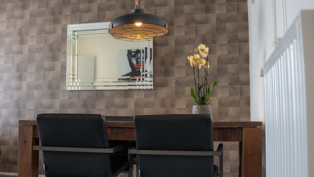 A dining area with a wooden table and two black chairs, a hanging pendant light, a mirror on the wall, and an orchid plant in a textured pot.