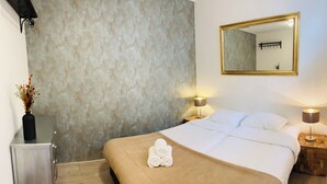 A hotel room with a bed, a wooden nightstand, a lamp, a mirror, and a vase with flowers.