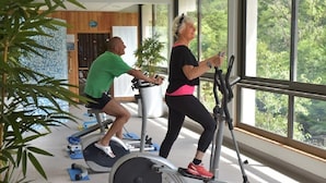 Two people exercising on stationary bikes in a room with large windows and a view of greenery outside. - LA CABANE AUX ECREVISSES- FEET IN THE WATER (Châteauneuf-les-Bains)