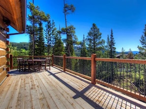 A wooden deck with a table and chairs, surrounded by a metal railing, overlooking a forested landscape.