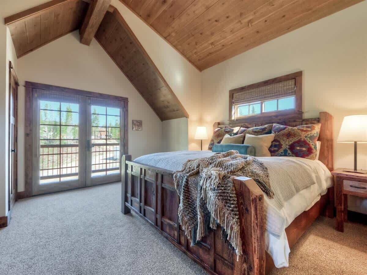 A bedroom with a sloped ceiling, a bed with a wooden headboard, a nightstand, a lamp, and a window with a view of greenery.