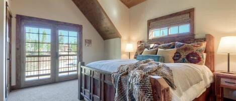 A bedroom with a sloped ceiling, a bed with a wooden headboard, a nightstand, a lamp, and a window with a view of greenery.
