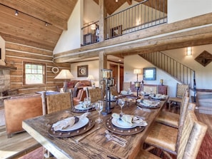 Un comedor amplio con una larga mesa de madera dispuesta para una comida, una zona de estar acogedora con sillas acolchadas, y una escalera que conduce a un nivel superior.