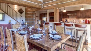 A dining area with a wooden table set for six, surrounded by wicker chairs. A kitchen with wooden cabinets and a bar area is visible in the background. - Luxury 5 Bedroom Big Sky Retreat (Big Sky)