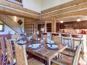 A dining area with a wooden table set for six, surrounded by wicker chairs. A kitchen with wooden cabinets and a bar area is visible in the background. - Luxury 5 Bedroom Big Sky Retreat (Big Sky)