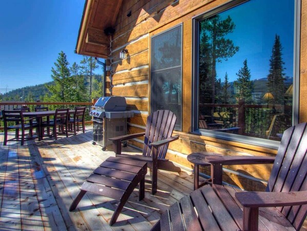 Une terrasse en bois avec des fauteuils Adirondack et un barbecue, donnant sur une zone boisée.