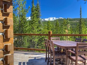 A wooden deck with a table and chairs, overlooking a forest and snow-capped mountains. - Luxury 5 Bedroom Big Sky Retreat (Big Sky)