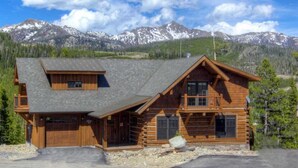 A log cabin with a steeply pitched roof, a front porch, and a garage. It is surrounded by a gravel driveway and a forested area with mountains in the background. - Luxury 5 Bedroom Big Sky Retreat (Big Sky)